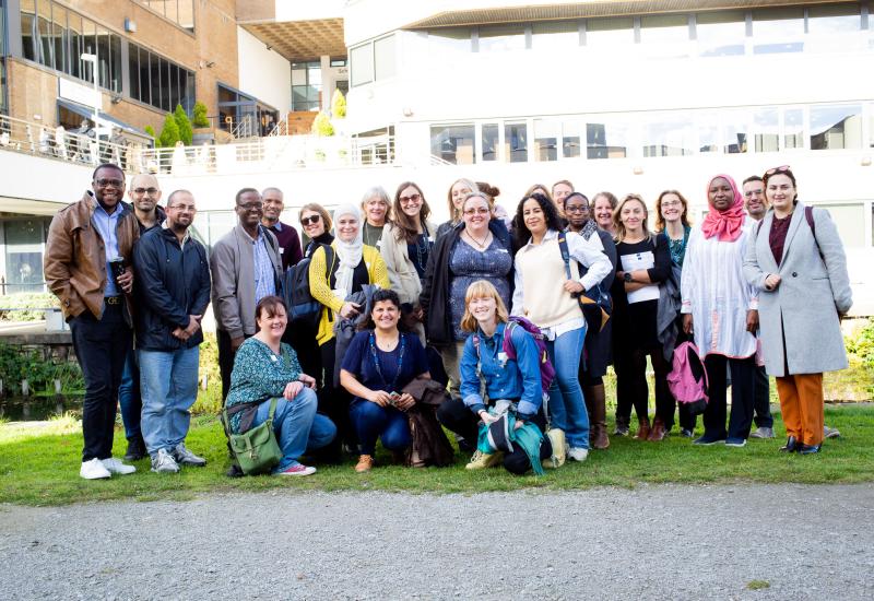 Photo of a group of about 25 people, standing or sitting on grass in front of a modern building.